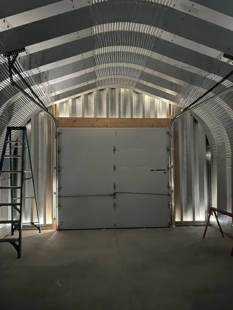 Interior of a metal building with a closed garage door, lit by overhead lights, with a ladder on the left.