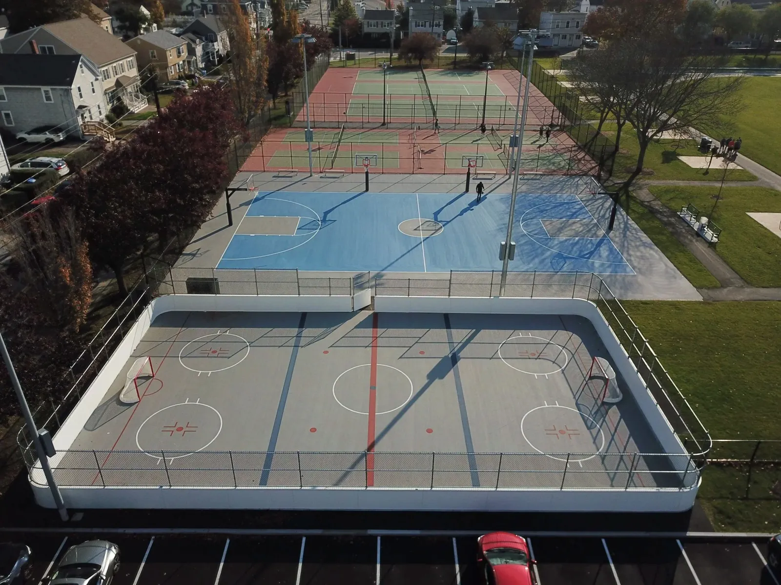 An aerial view of multiple sports courts in a park, including an ice hockey rink, a basketball court, and tennis courts, surrounded by trees, residential houses, and a parking lot with cars.