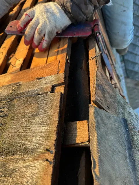 Gloved hand using a knife to pry up roof material. Wood framing is visible.