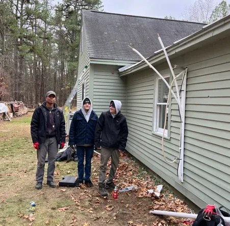 Three people standing beside a house with damaged siding and roof, debris scattered on the ground.