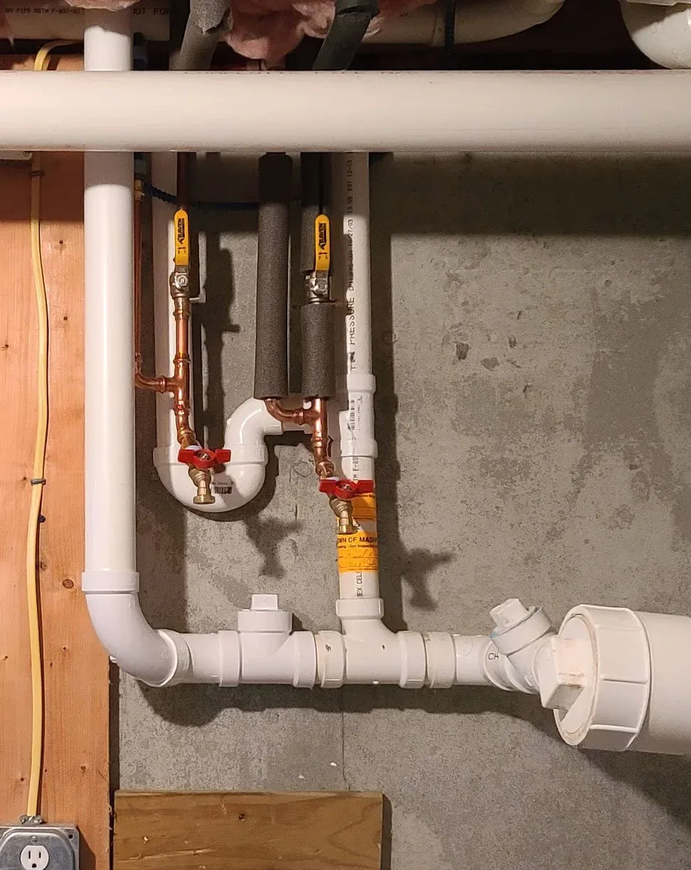 Plumbing pipes with shut-off valves in a utility room, including a white PVC drain pipe, copper cold and hot water supply lines, and two yellow-handled valves.