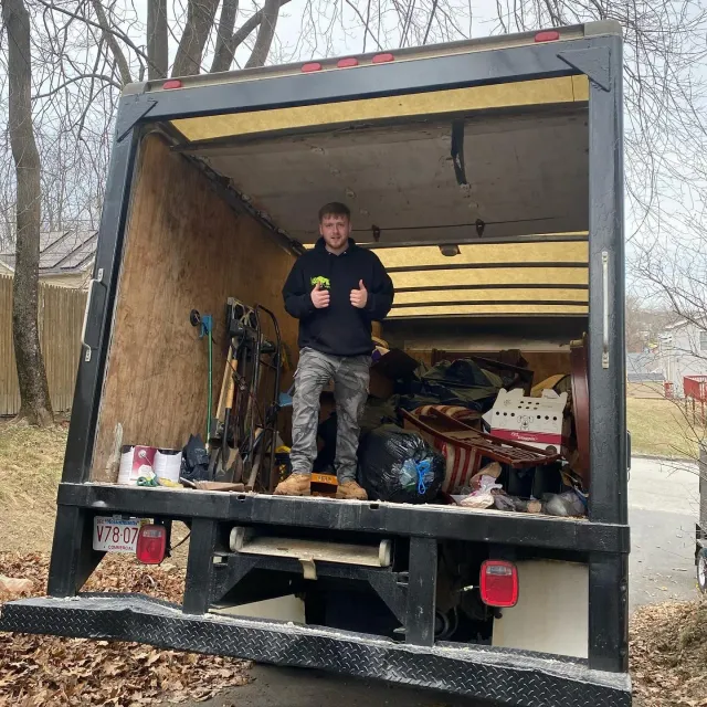 Man standing in the back of a moving truck with thumbs up, surrounded by items. Outdoor setting.