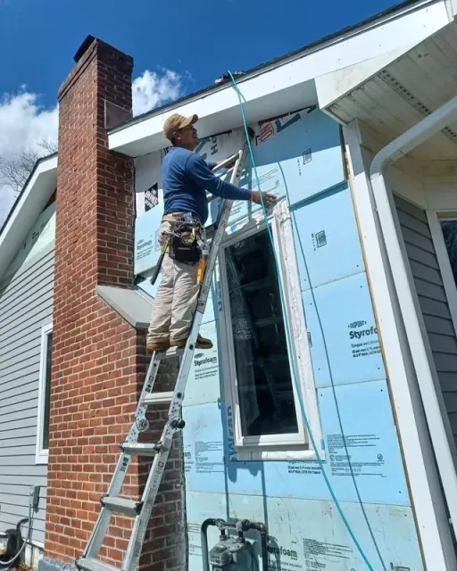 Construction worker on ladder installing a window on a house with blue insulation. Chimney is visible.