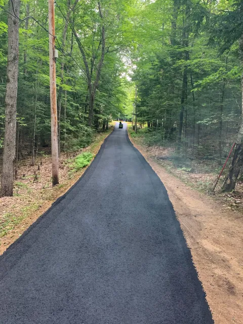 A newly paved narrow asphalt road winding through a lush green forest, with wooden utility poles and power lines on the left side and trees on both sides.