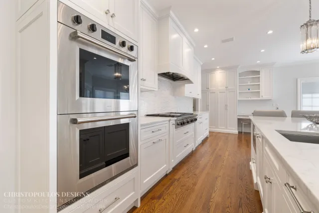 Modern white kitchen with stainless steel double oven, white cabinets, marble countertops, and wood flooring.