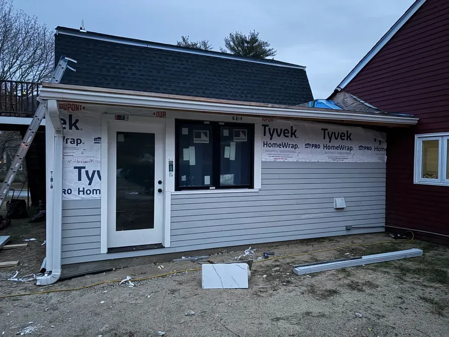 A single-story home addition with gray horizontal siding, black-framed windows, and exposed Tyvek house wrap.