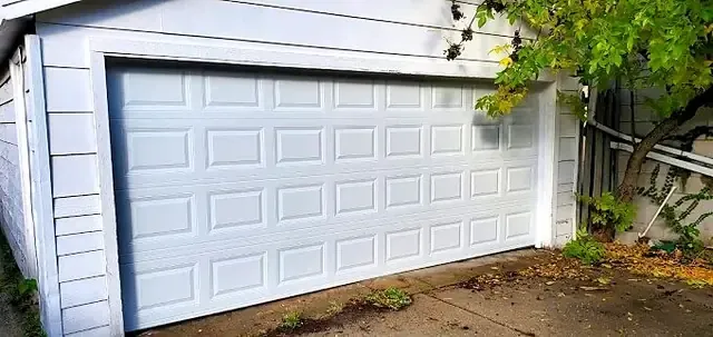 White raised-panel double garage door on a gray-sided garage with a tree branch overhead