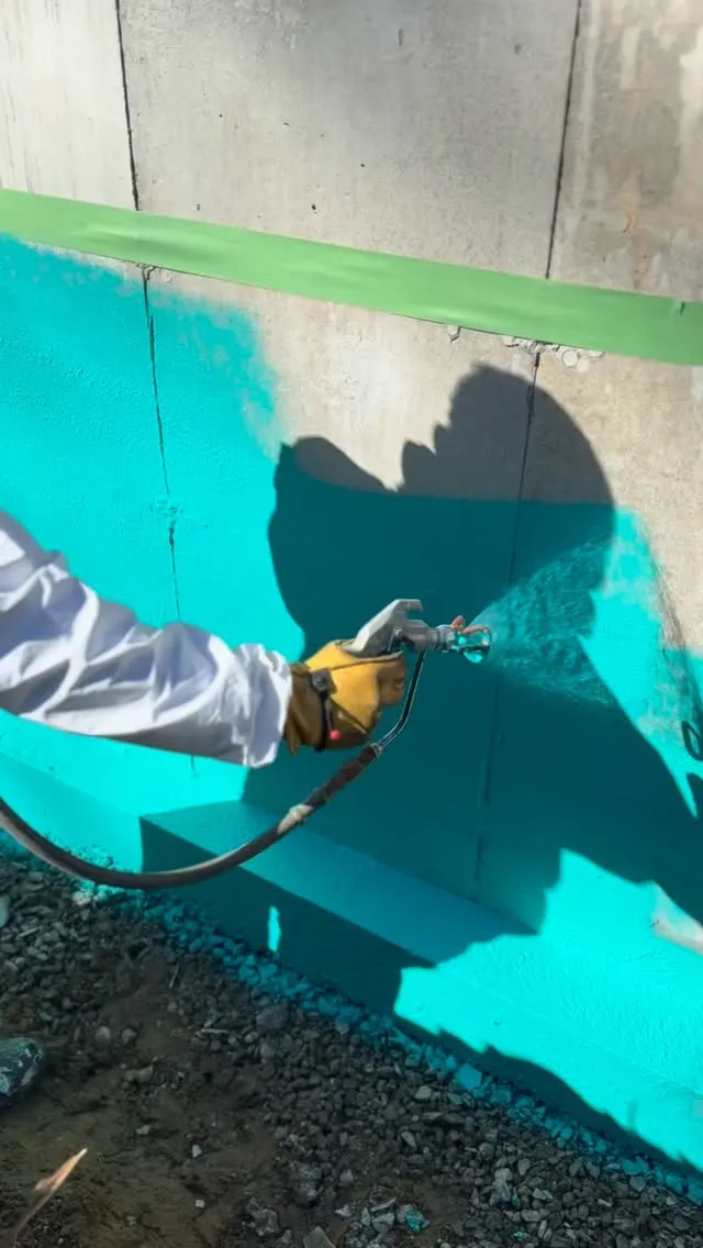 Crew member spray-applying a teal waterproofing membrane to a foundation wall
