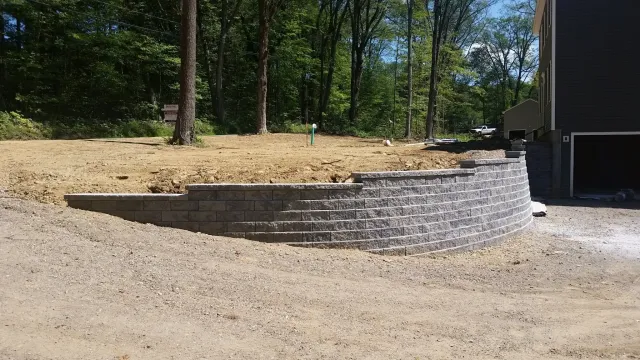 A newly built gray stone retaining wall with a curved design in front of a house, with an area of bare earth behind it, surrounded by trees and a wooded area in the background.