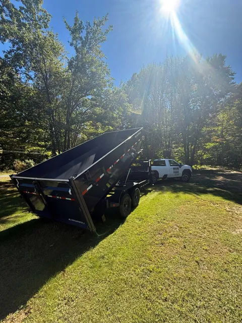 Dump trailer tilted up, being towed by a white truck on green grass with sun shining through trees.