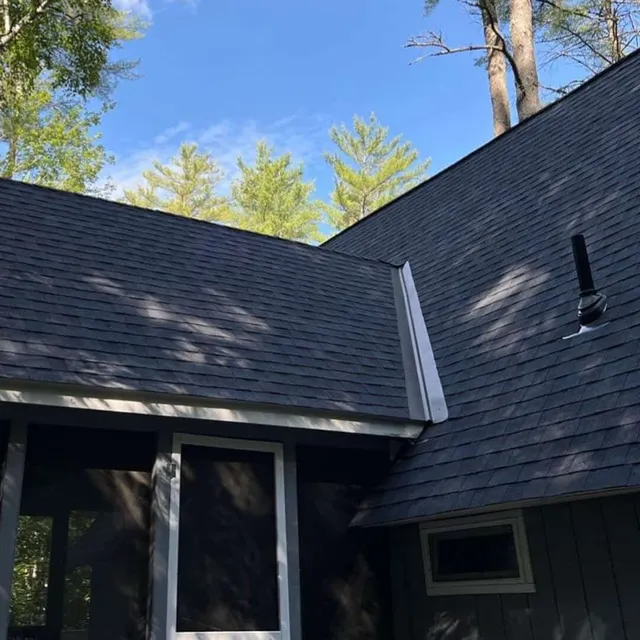 A house with dark gray shingled roofs, surrounded by trees under a clear blue sky.