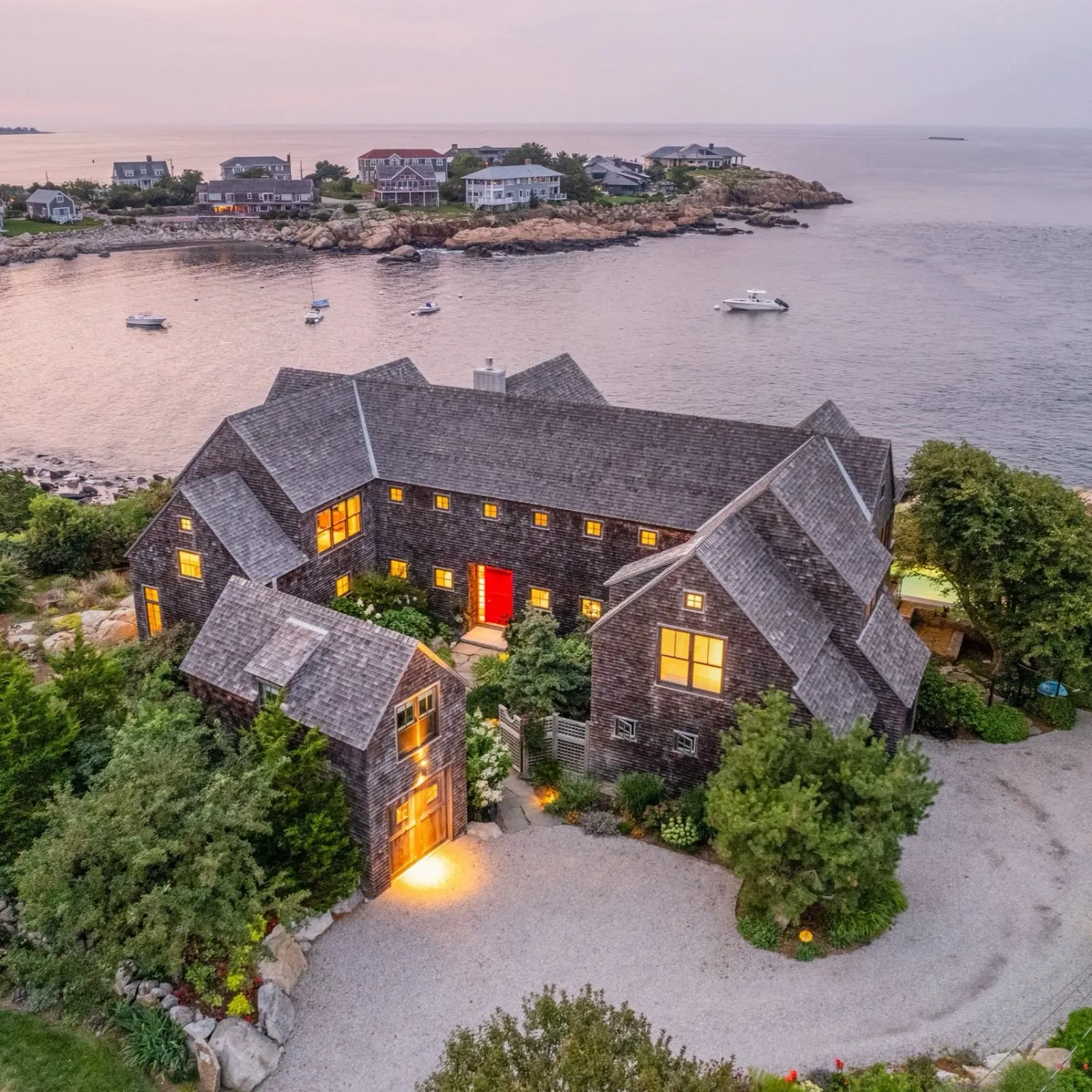 Modern shingle-style home with a red door at sunset, overlooking the ocean on Cape Ann with boats and distant shoreline views.
