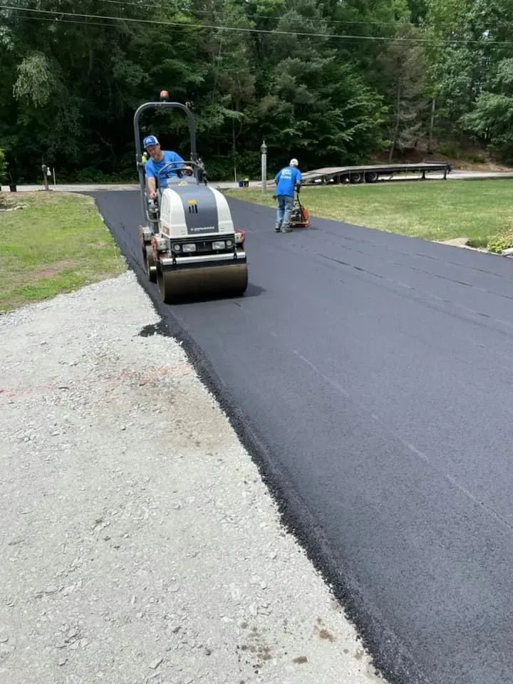 Two workers using a construction roller to smooth fresh asphalt on a driveway next to a gravel area.