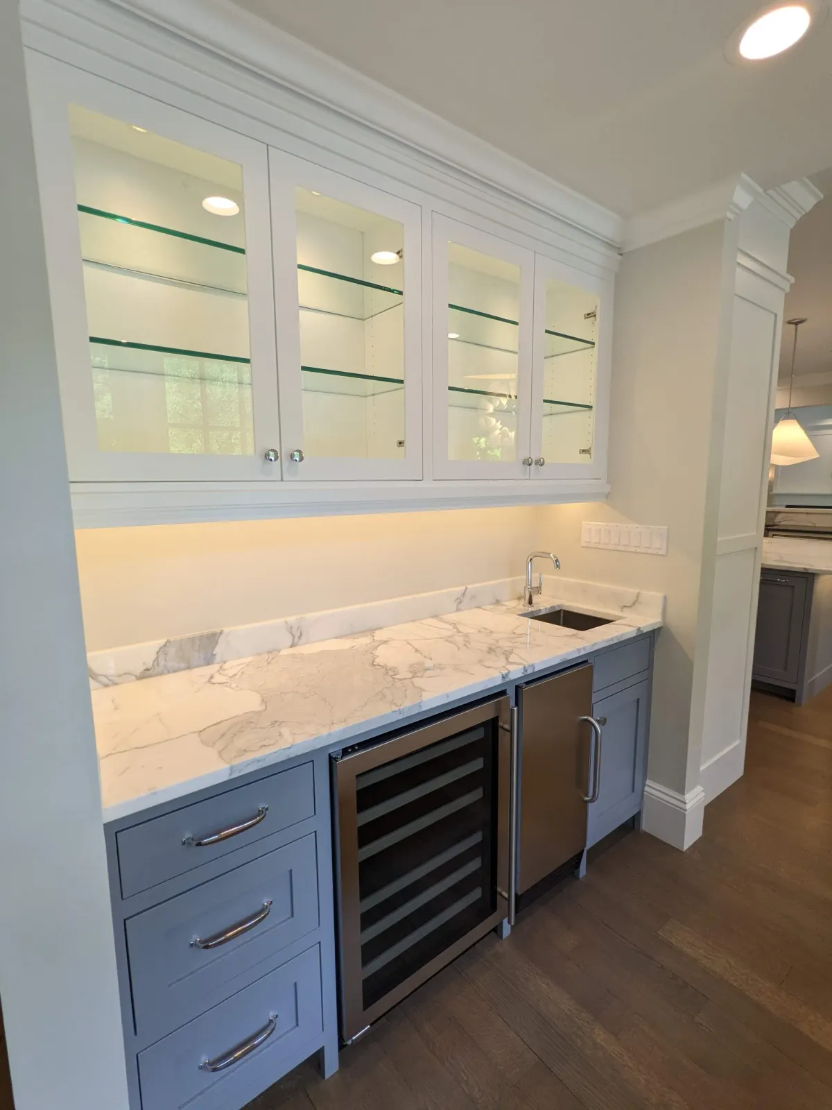 A kitchen wet bar area with gray cabinets, a marble countertop, a small stainless steel refrigerator, a built-in wine cooler, and upper glass-front cabinets with glass shelves, under-cabinet lighting, and a small sink.