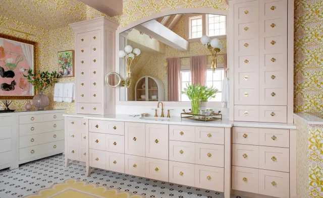 Elegant bathroom with floral wallpaper, pink cabinets, and brass fixtures. Large mirror with twin globe lights, decorative vases, and tulips on the countertop. Patterned floor tiles and framed artwork add accents.