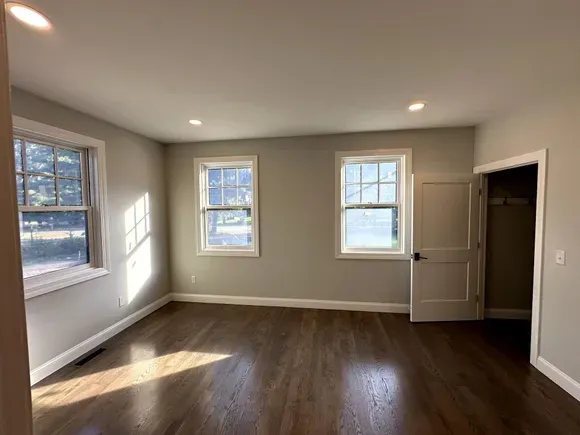 Empty beige room with hardwood floors, three windows, and an open door letting in sunlight.