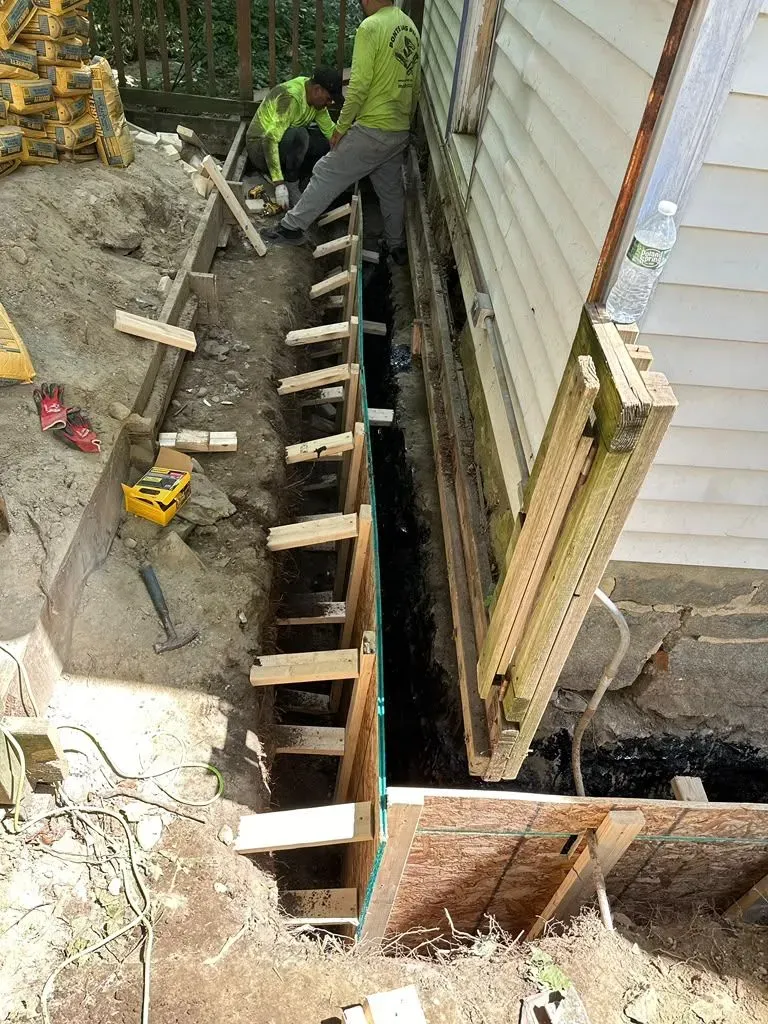 Workers building a concrete foundation beside a house, with wooden forms and a trench