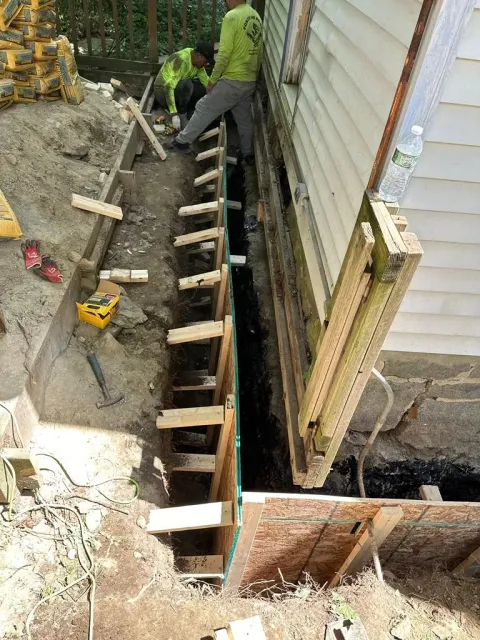 Workers building a concrete foundation beside a house, with wooden forms and a trench
