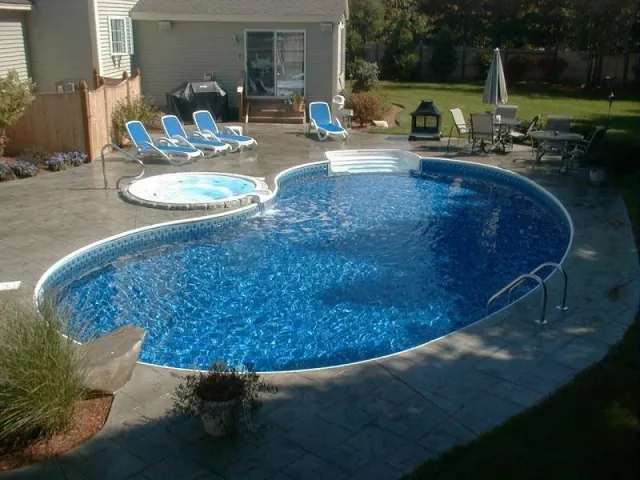 Backyard with a kidney-shaped swimming pool, a hot tub, lounge chairs, outdoor dining table with chairs, a grill, and a grassy yard with trees.