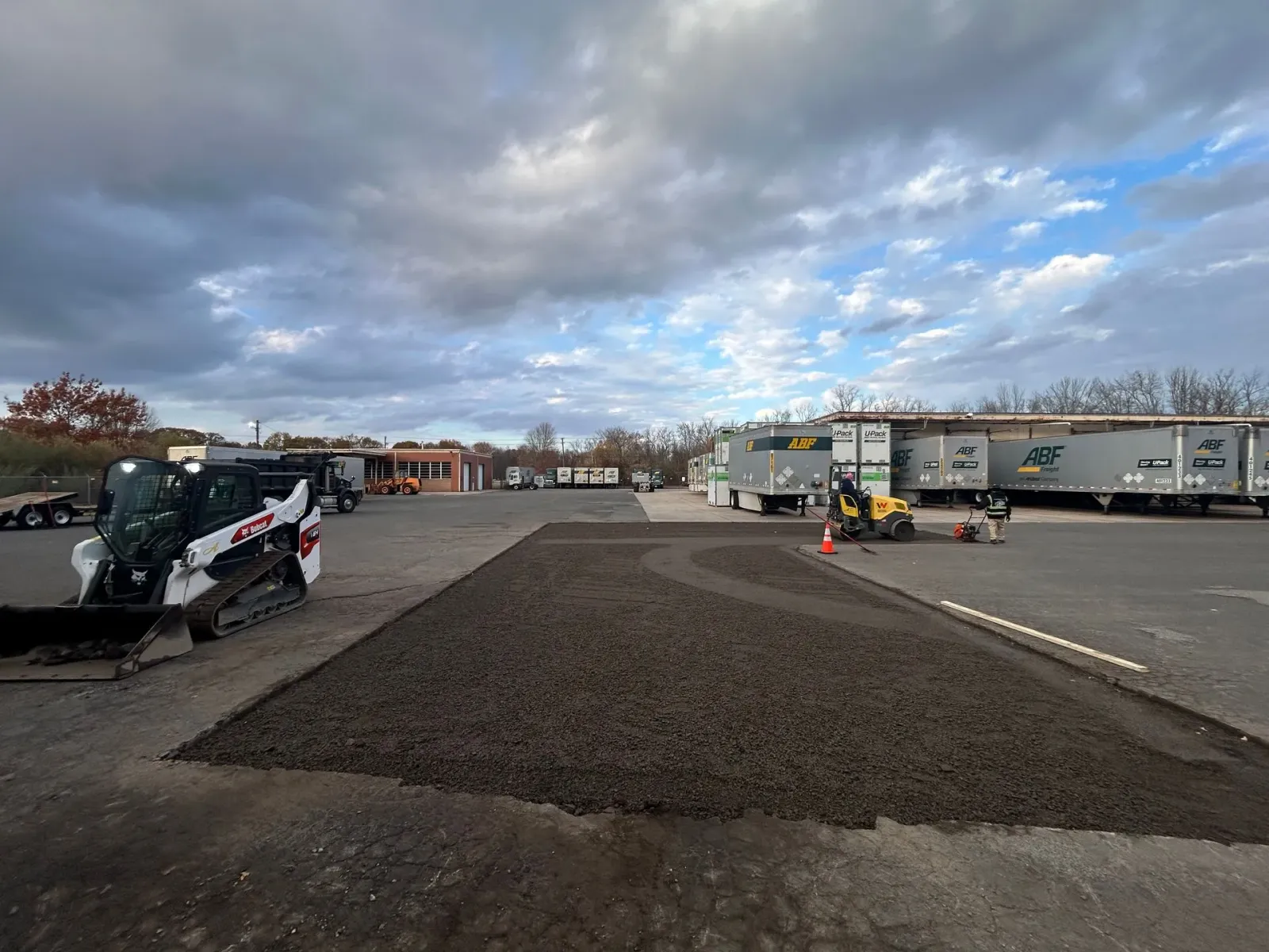 A construction site with a Bobcat loader parked beside a freshly laid patch of gravel against a cloudy sky.