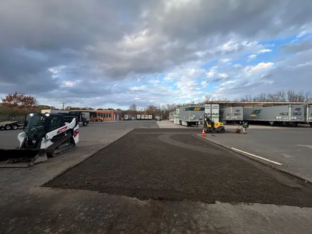 A construction site with a Bobcat loader parked beside a freshly laid patch of gravel against a cloudy sky.