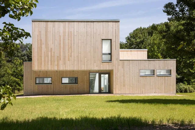 A modern two-story house with a wooden exterior, multiple windows, and a flat roof, surrounded by green grass and trees.