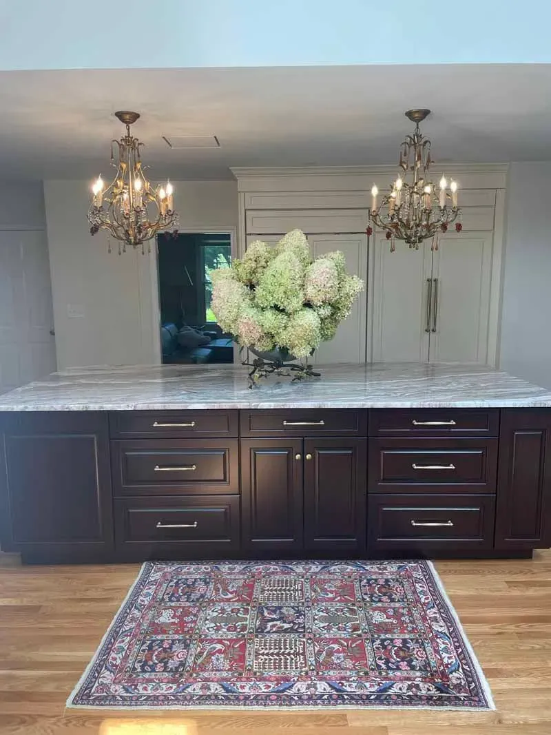Kitchen island with dark cabinetry, marble countertop, and chandeliers. Hydrangeas sit on the island, and a rug on the hardwood floor.