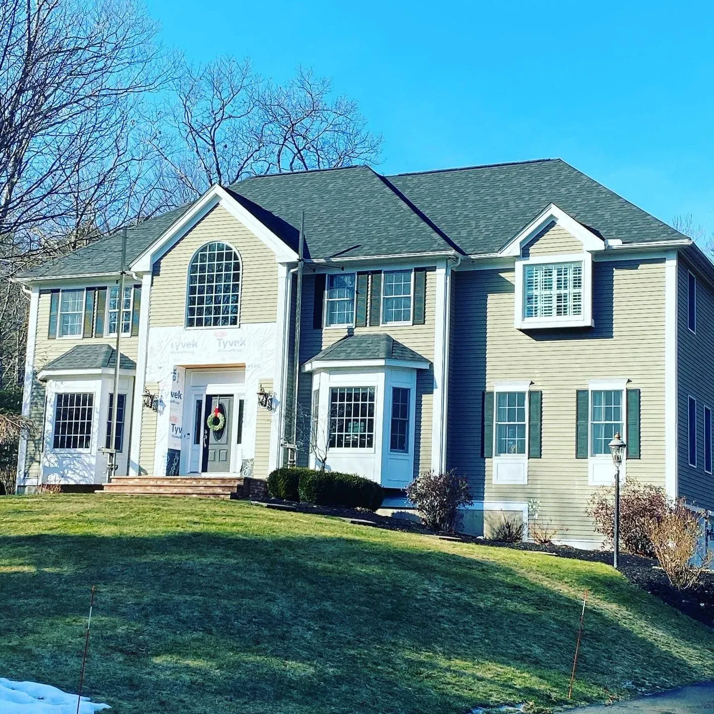 A large two-story house with beige siding, green shutters, and a dark gray roof, decorated with a holiday wreath, set against a clear blue sky and bare trees.