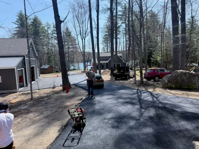 People are paving a driveway near a lake in a wooded area, with construction equipment and parked cars around.