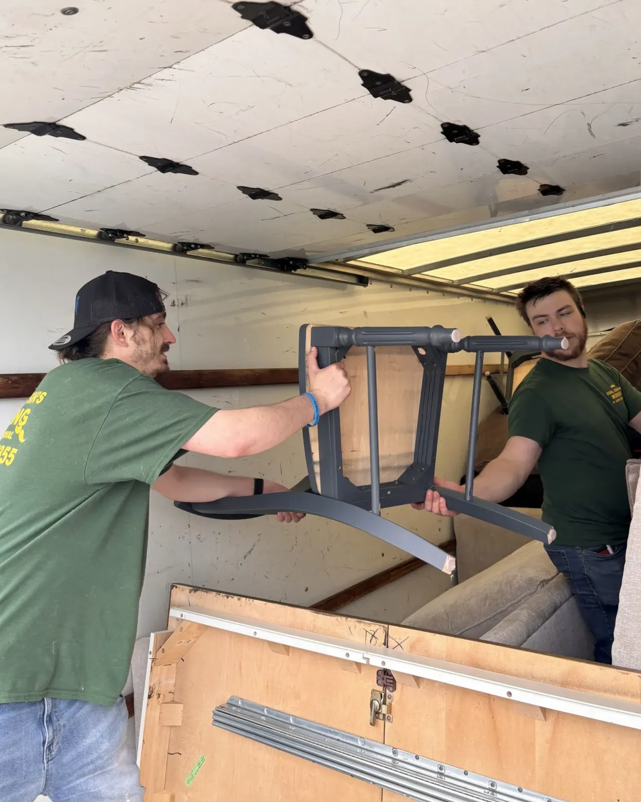 Two people lifting a dark framed chair inside a truck or moving van