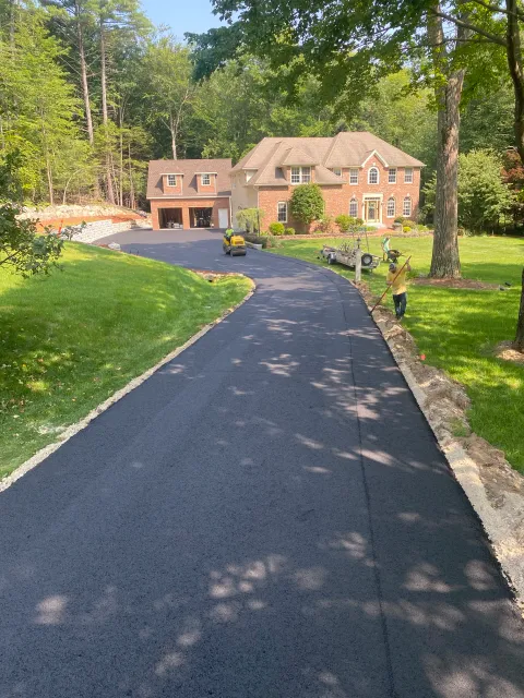 Newly paved driveway leading to a large brick house with a two-car garage. Two workers are working on the edges of the driveway, and landscaping equipment is present. The house is surrounded by lush green trees and grass.