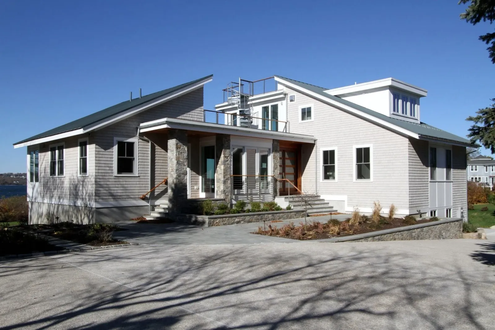 A modern multi-story house with light-colored siding, large windows, and a flat roof with a spiral staircase on the roof deck. There is a small front porch with stone columns and a landscaped yard with mulch and plants. An empty parking lot is in front of the house with shadows of trees on the pavement. The sky is clear and blue.