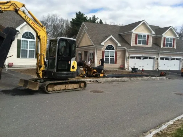Construction site with a mini-excavator removing pavement in front of beige houses with red shutters.