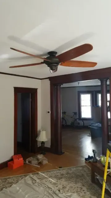 Ceiling fan in a living room with dark wood trim, leading to a second room with bicycles and hardwood floors.