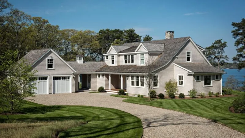 Large light-colored house with gray roof by a curved driveway, grass, trees, and water – McPhee Associates of Cape Cod.