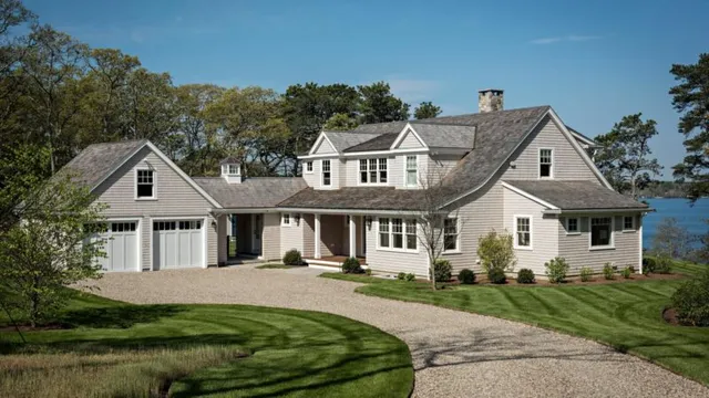 Large light-colored house with gray roof by a curved driveway, grass, trees, and water – McPhee Associates of Cape Cod.