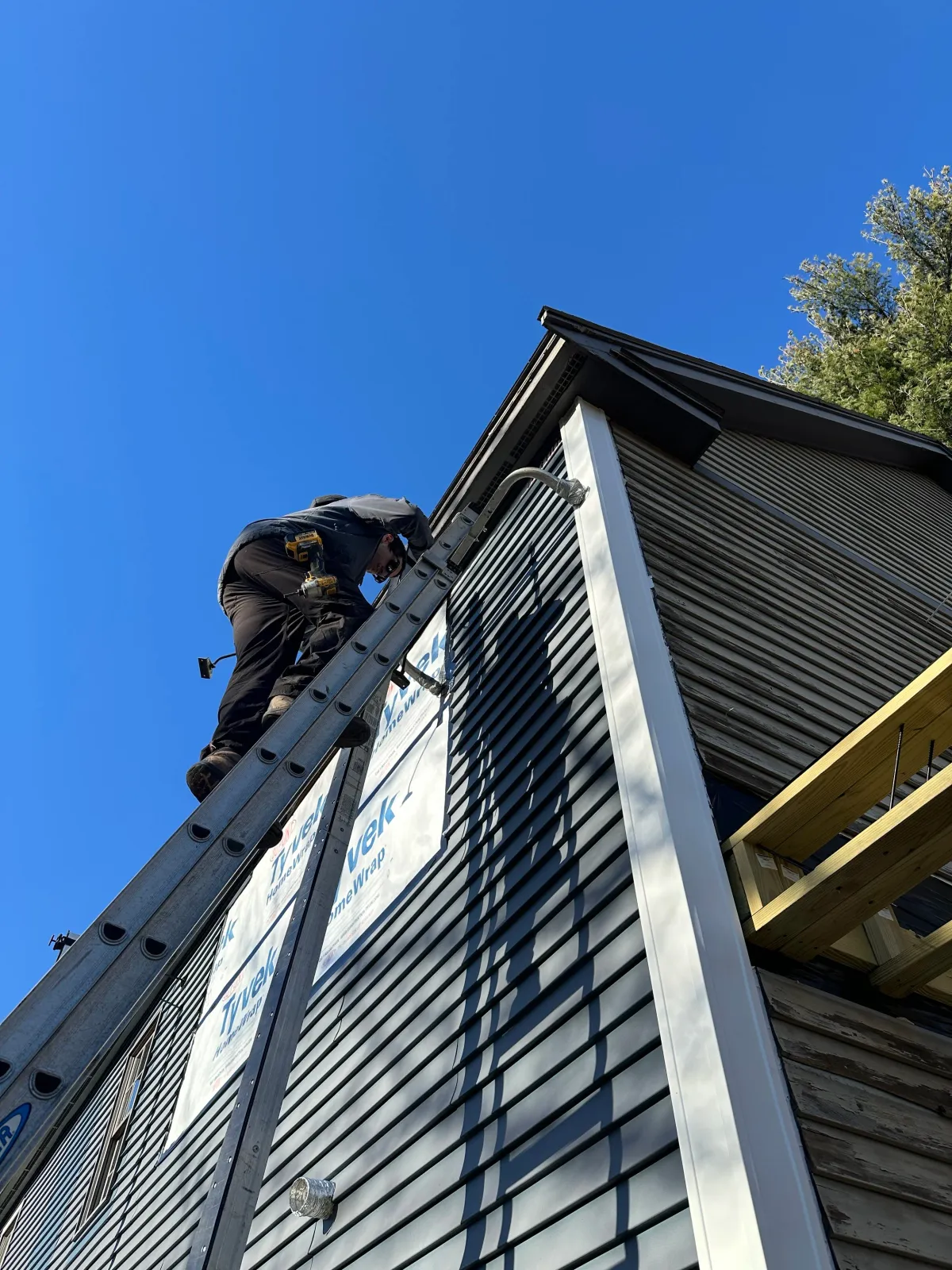 Worker on a ladder installing or repairing siding on the upper side of a house under a clear blue sky.