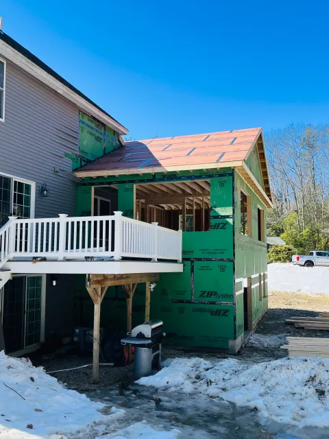 Construction site of an addition to a house, with green insulated sheathing and a new roof being built, snow on the ground, and construction materials nearby.