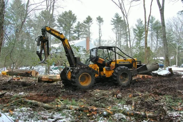 A yellow compact excavator with the brand name 'John Deere' on its side, working on a wooded area, with logs and debris around it, and some snow on the ground.