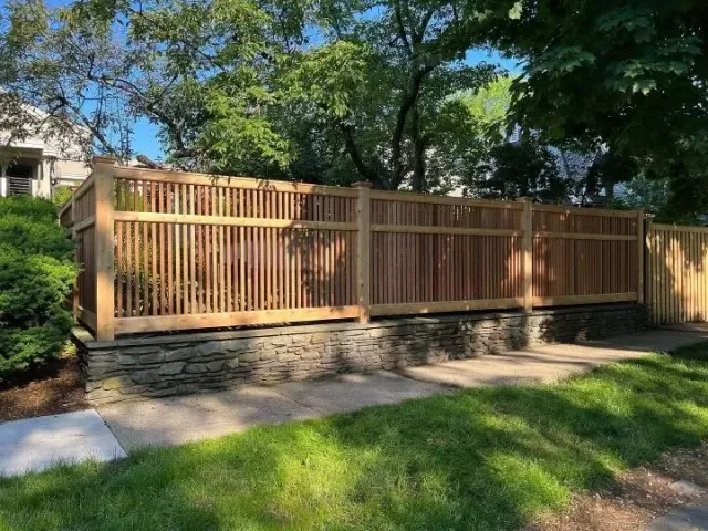 Wooden privacy fence with stone base along a sunny residential sidewalk and green lawn