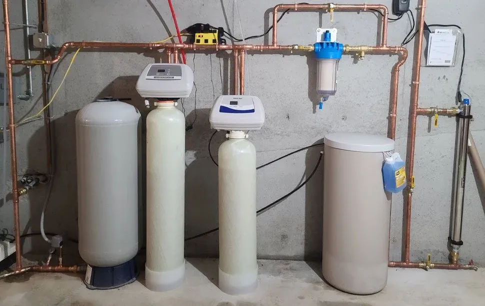 Water filtration system with various tanks, control valves, and copper piping against a concrete wall.