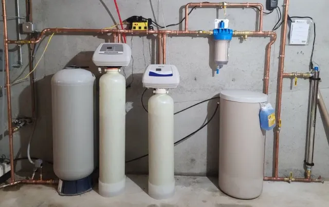 Water filtration system with various tanks, control valves, and copper piping against a concrete wall.