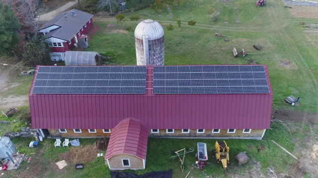 Aerial view of a farm building with a red metal roof covered with solar panels; in the background, a white grain silo, a small red house with a gray roof, and farmland with some equipment and trees.