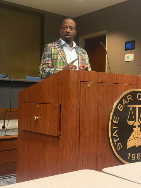 Man speaking at a wooden podium in a courtroom, wearing a colorful plaid jacket.