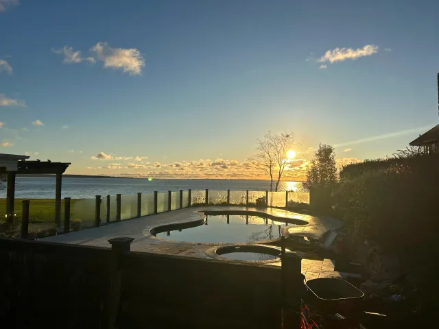 Sunset view over a body of water with a backyard pool in the foreground, surrounded by a wooden fence and trees.