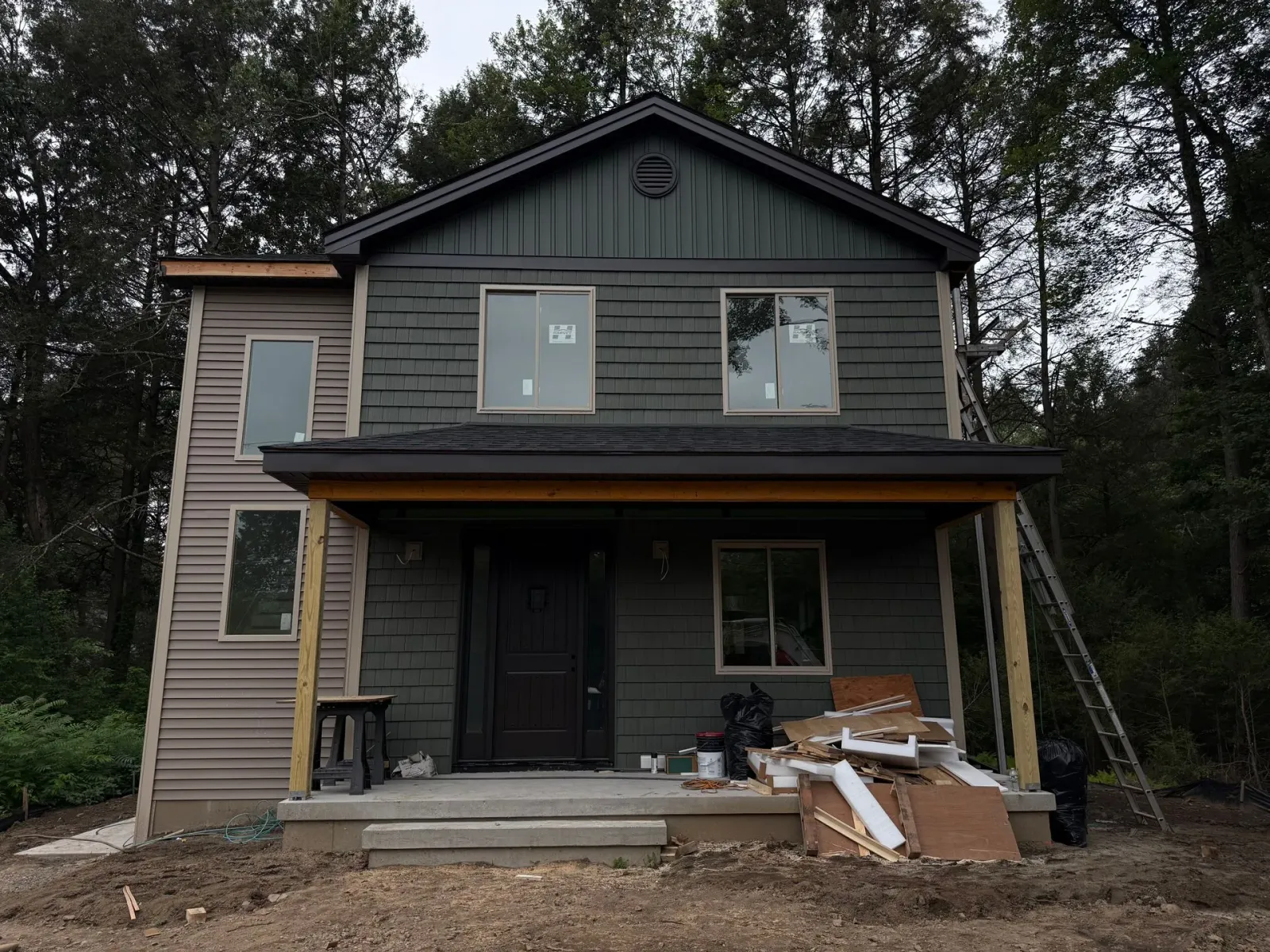 Two-story house with dark green siding, covered porch, and construction materials scattered on the porch.