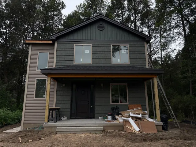 Two-story house with dark green siding, covered porch, and construction materials scattered on the porch.
