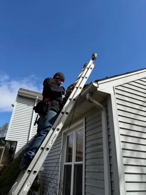 Worker on extension ladder inspecting roof edge of a gray house under a blue sky