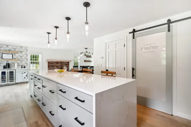 Bright, modern kitchen with large island, pantry door, pendant lights, and wooden floors.