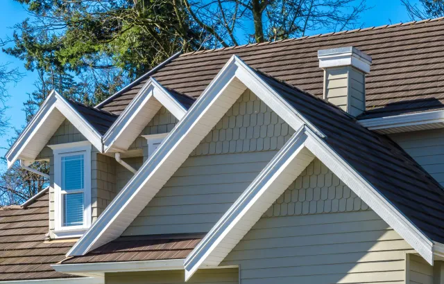 A residential roof with multiple gables, white trim, light-colored wood-shingle siding, and a brick chimney.
